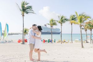Couple sharing a romantic kiss on a tropical beach with palm trees and a cruise ship in the background.