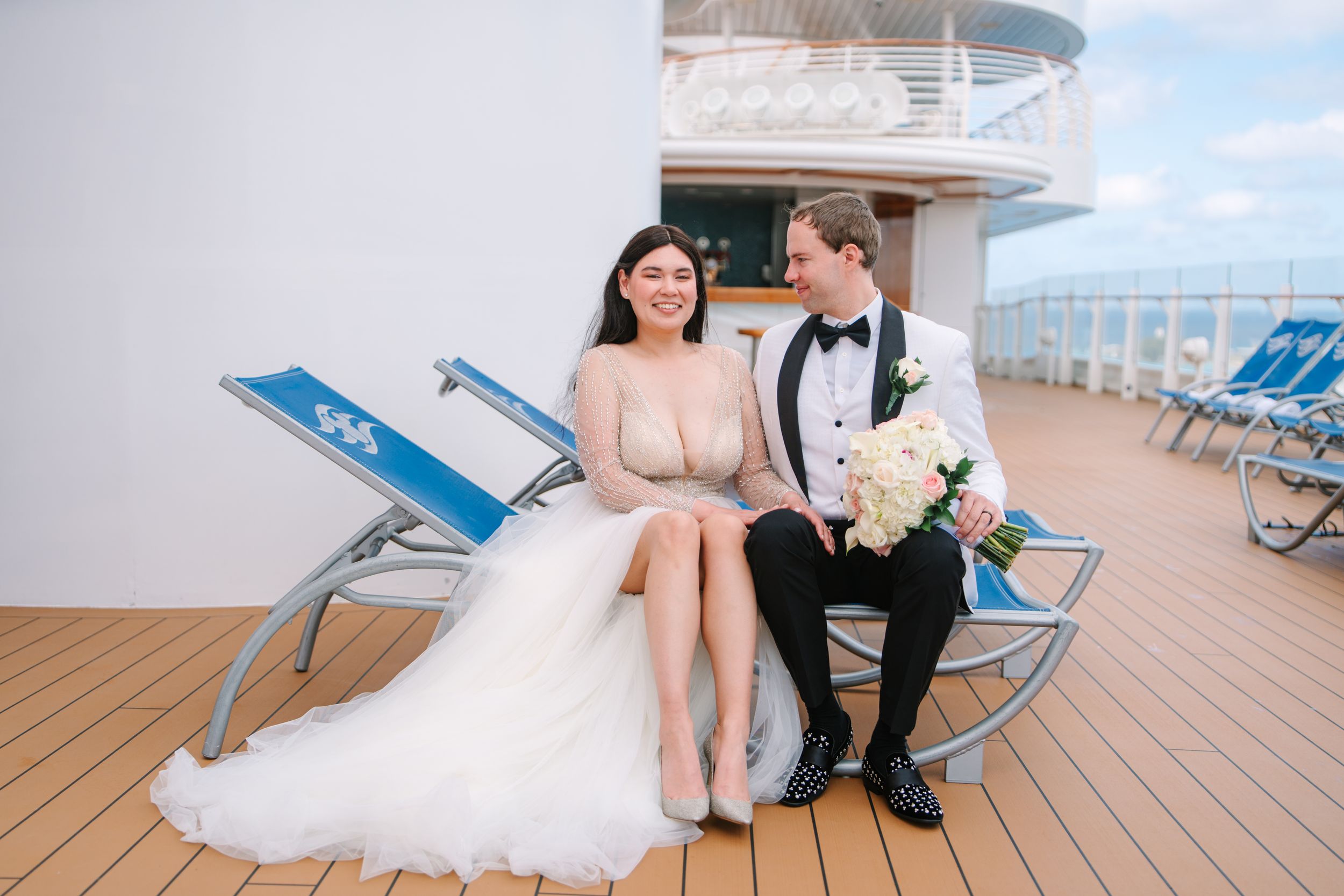 Bride and groom in wedding attire sitting on deck chairs aboard a luxury cruise ship on their wedding day.