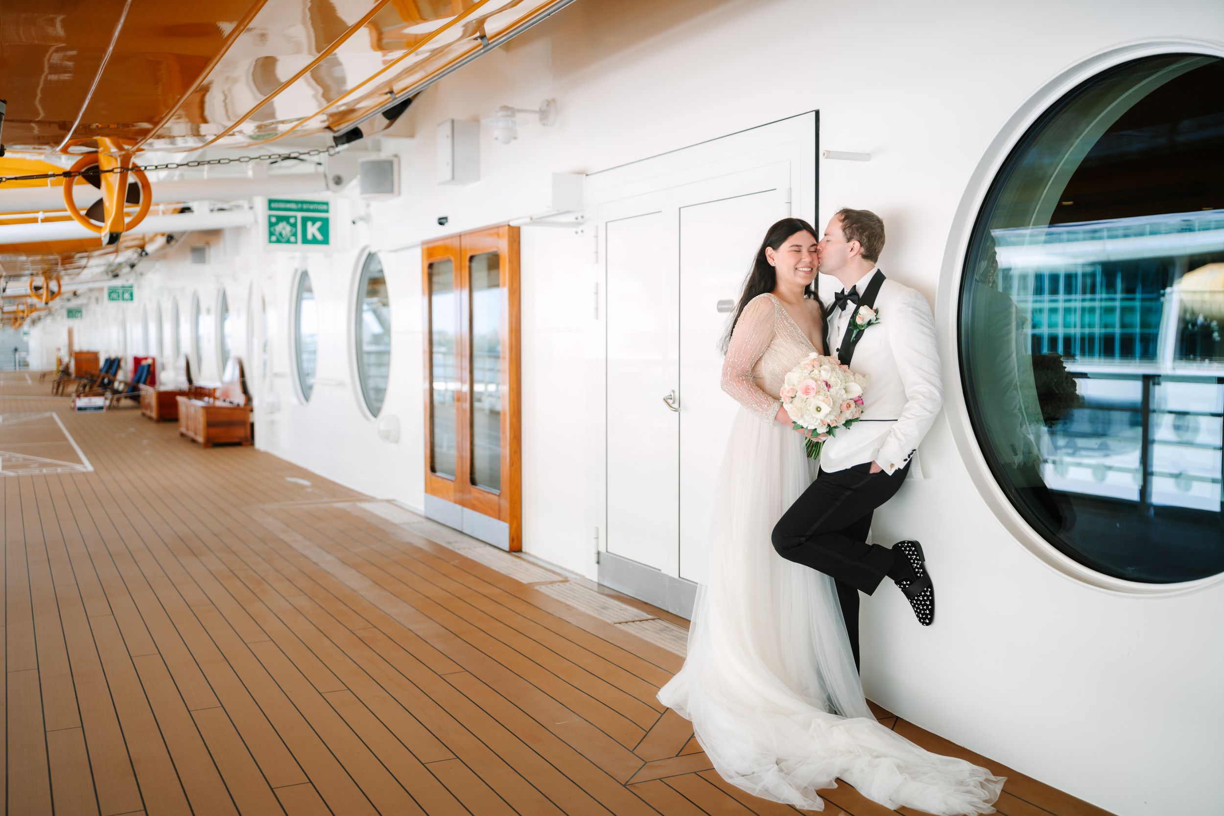 Bride and groom sharing a romantic kiss on a cruise ship deck, with the groom playfully jumping beside a porthole window.