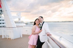 Couple embracing on cruise ship deck at sunset, woman in pink dress, man in white tuxedo jacket, ocean backdrop.