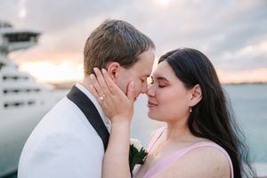 Couple sharing a romantic almost-kiss at sunset near a cruise ship, bride in pink dress touching groom's face.