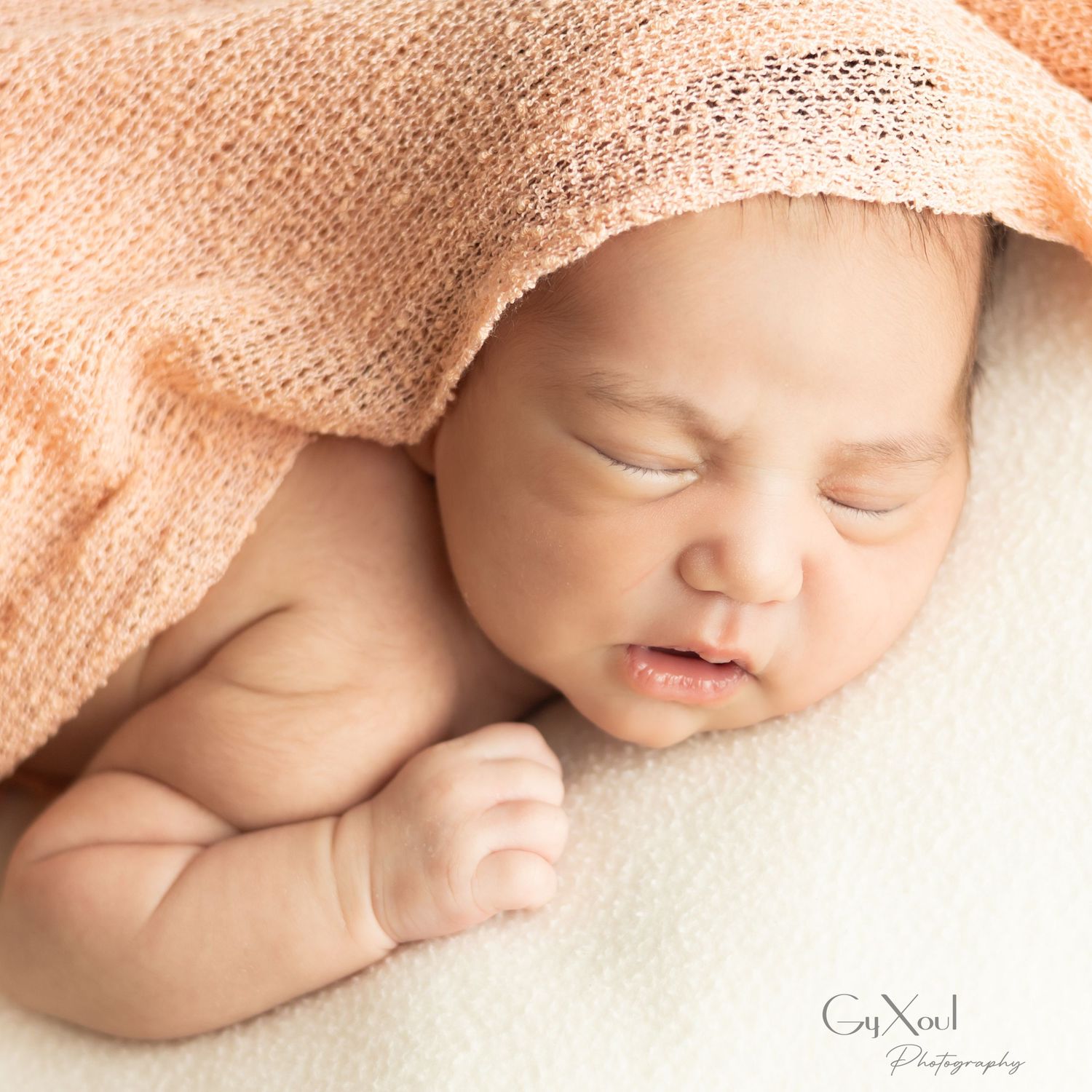 Peaceful newborn baby sleeping under a soft peach knit blanket in a professional photography portrait.