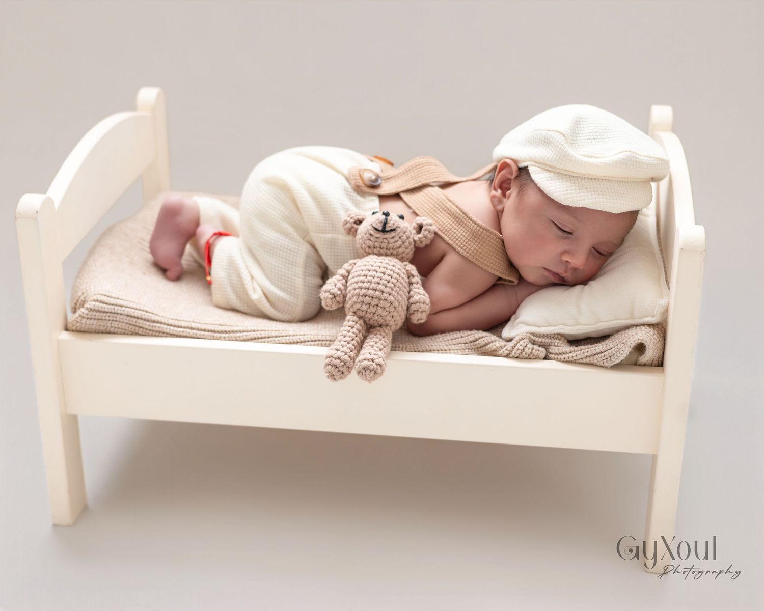 Newborn baby sleeping peacefully on a tiny white wooden bed with a crocheted teddy bear, wearing a cream outfit and hat.