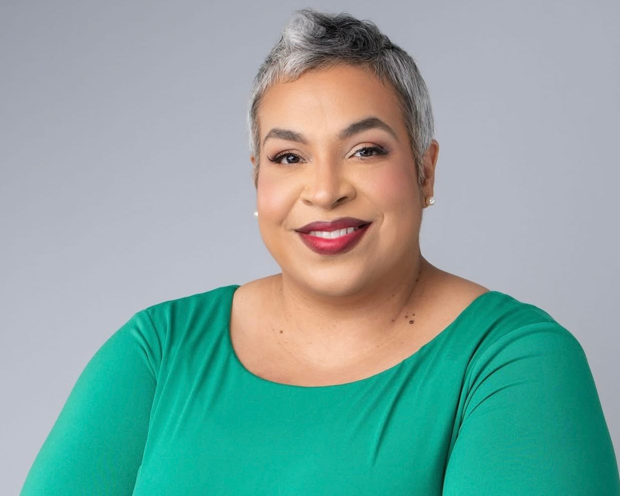Professional headshot of a person with silver hair wearing a royal blue v-neck top and bright red lipstick against a gray background.