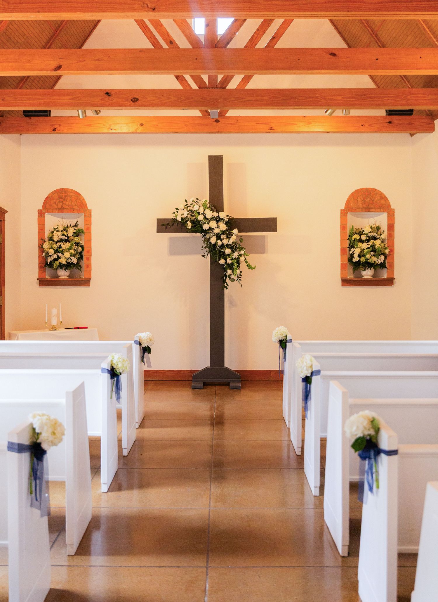 White church pews line the aisle of a chapel decorated with floral arrangements and a wooden cross for a wedding ceremony.