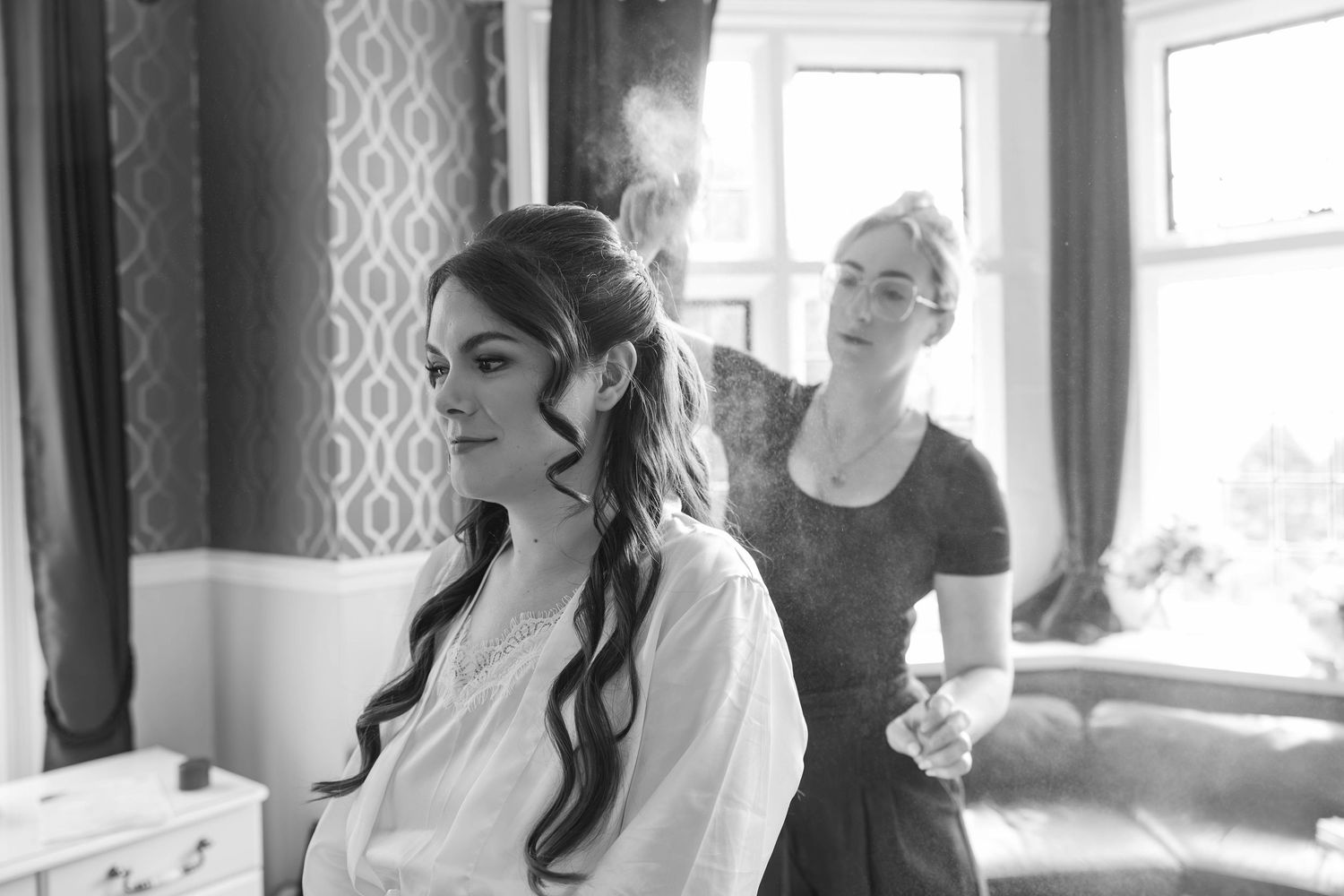Hair stylist applies finishing spray to brides elegant braided hairstyle in black and white bridal prep photo.