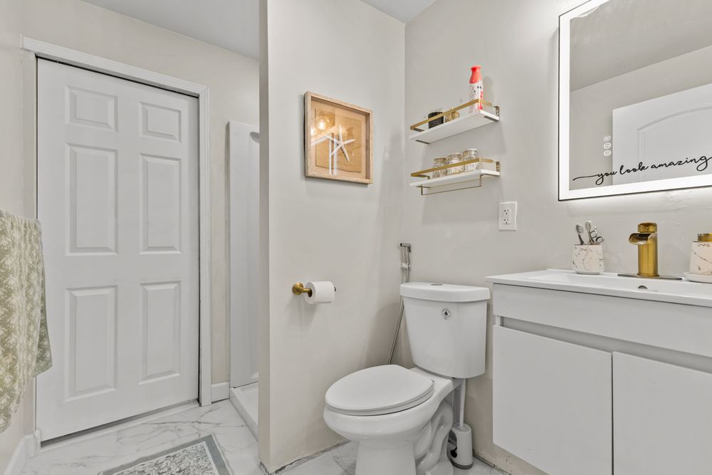 Clean white bathroom with toilet, vanity, illuminated mirror, open shelving, and tiled flooring.
