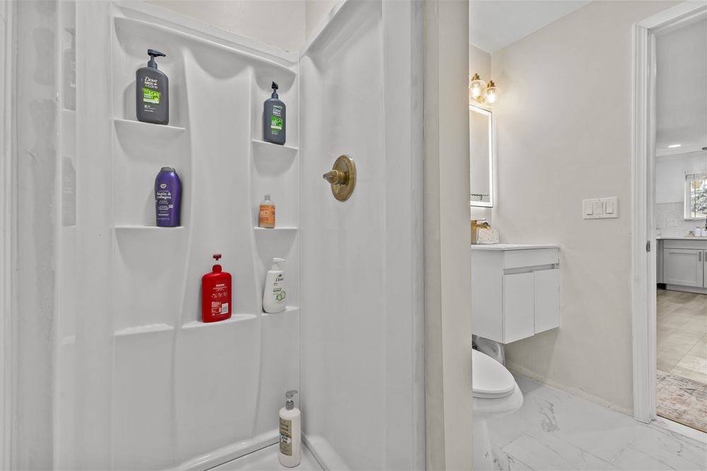 Clean white walk-in shower with built-in shelves and gold fixtures next to a modern vanity.