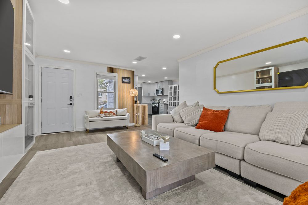 Bright living room with beige sectional sofa, orange accent pillows, gray coffee table, and open view to kitchen.