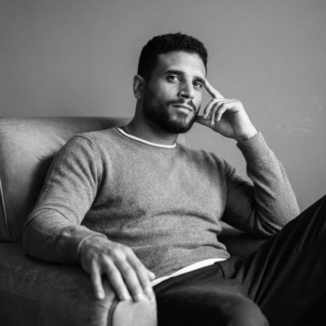 Black and white portrait of a young man in a sweater sitting relaxed in a leather chair, resting his chin on his hand.