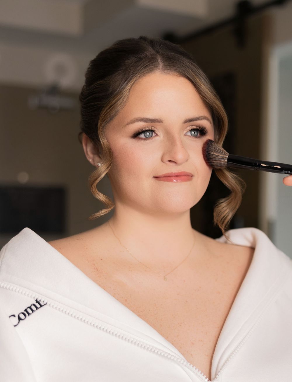 Bride getting makeup applied with a brush during wedding day preparations in a white robe.