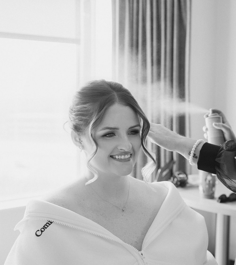 Black and white portrait of a smiling bride getting hair spray applied during wedding day preparations.