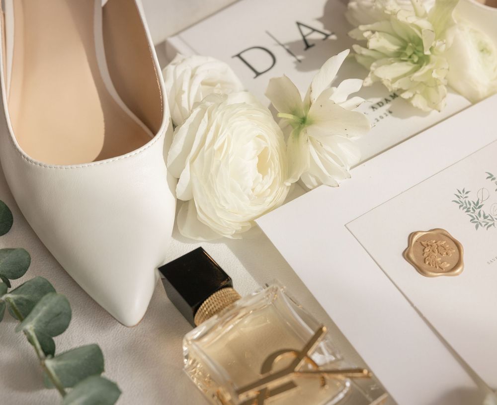 Bridal flat lay with white heels, ranunculus flowers, YSL perfume, and wax seal invitation on white background.