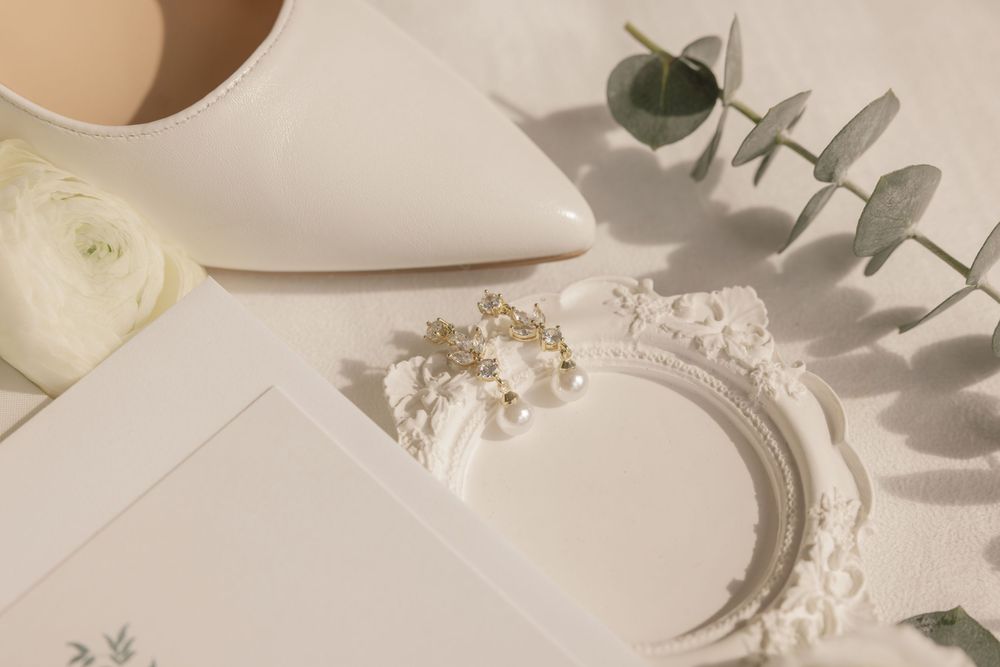 Elegant wedding flat lay with white heels, floral hair accessories, envelope, and eucalyptus on linen.