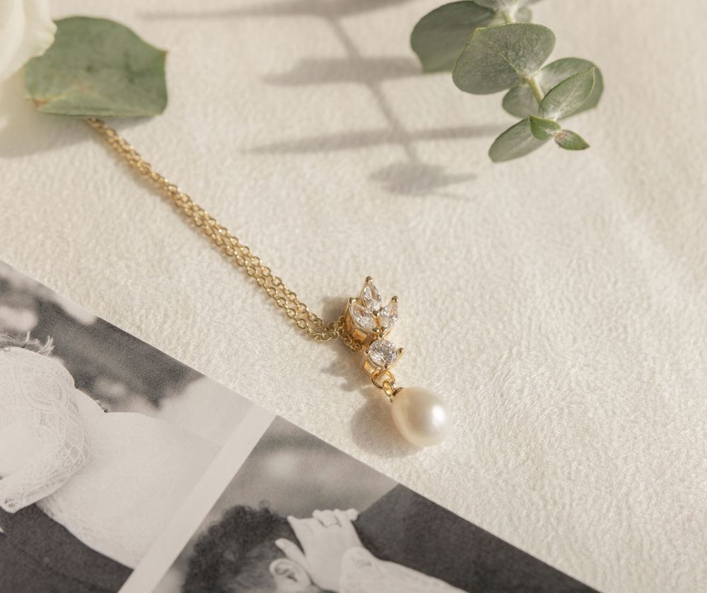 Elegant gold pearl and diamond drop necklace displayed on white fabric with eucalyptus leaves and wedding photos.