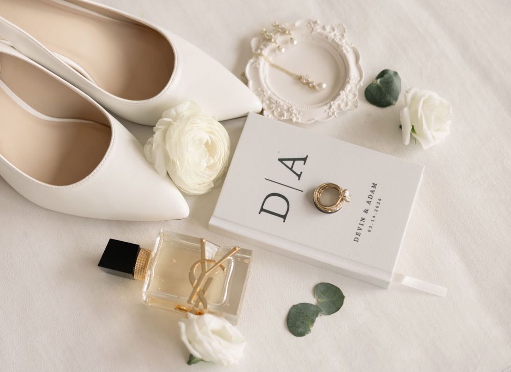 White wedding flat lays with heels, perfume, earrings, roses, and monogram card on white background. brookshire venue