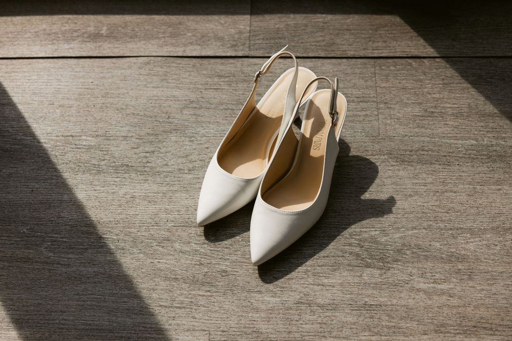 Elegant white pointed-toe slingback wedding heels resting on gray tile flooring in soft natural light.