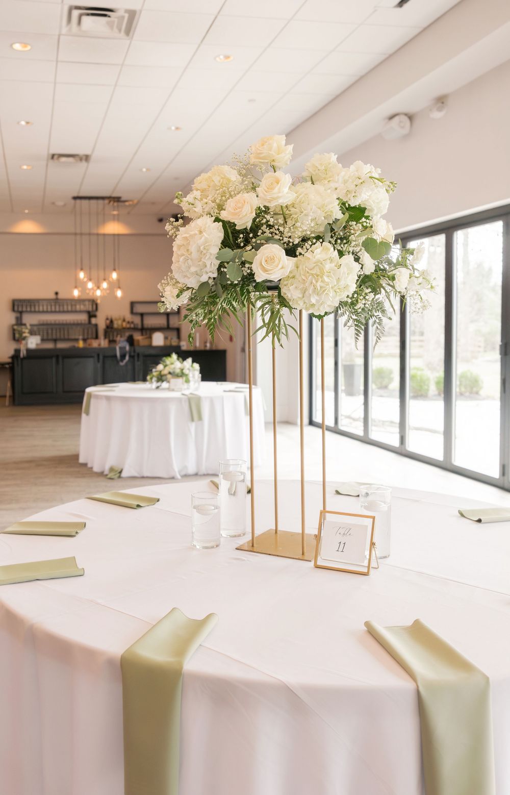 Elegant wedding reception table with tall gold floral centerpiece featuring white roses and hydrangeas, sage green napkins. Brookshire venue