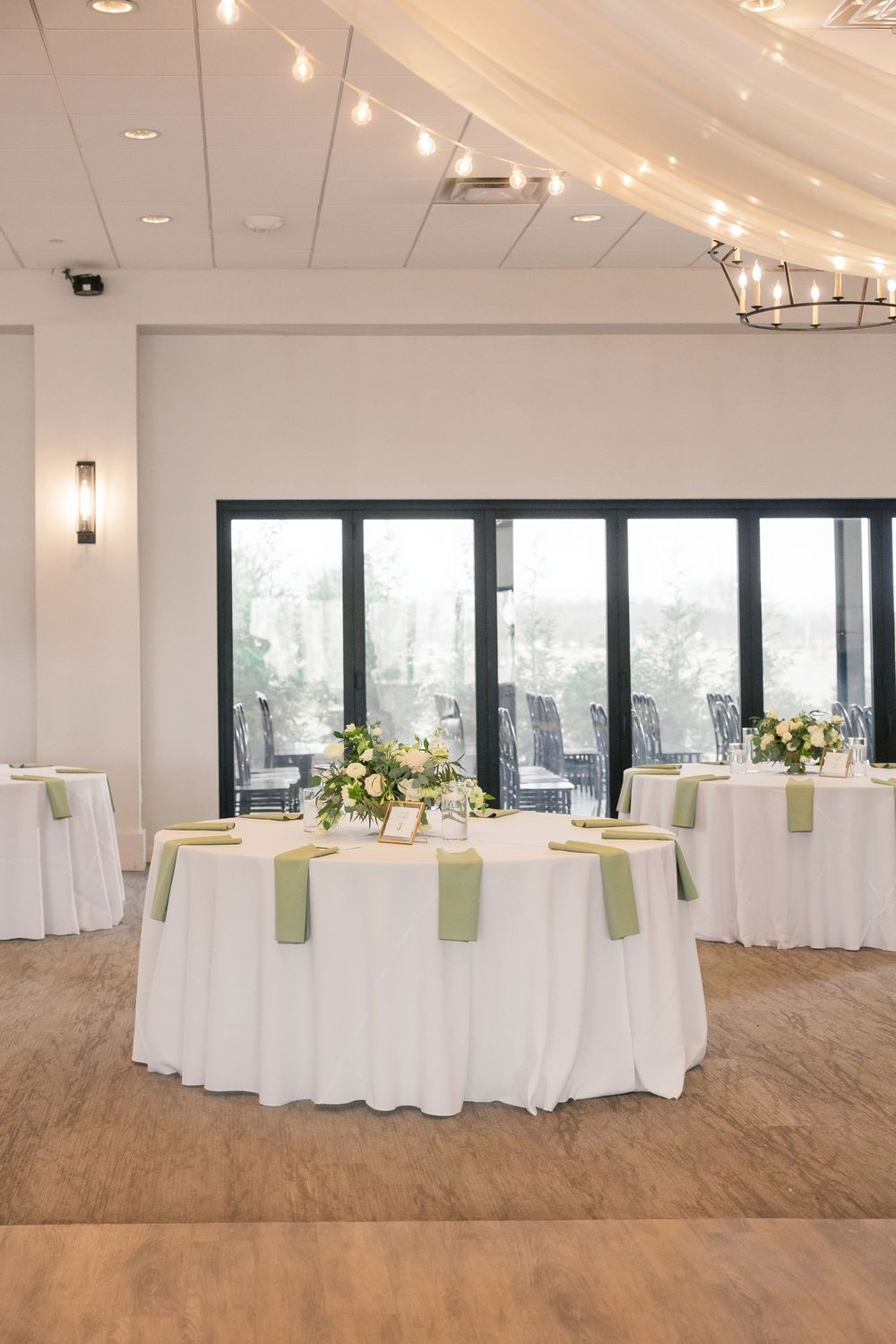 Elegant wedding reception hall with white tablecloths, sage green napkins, floral centerpieces, string lights, and large windows.