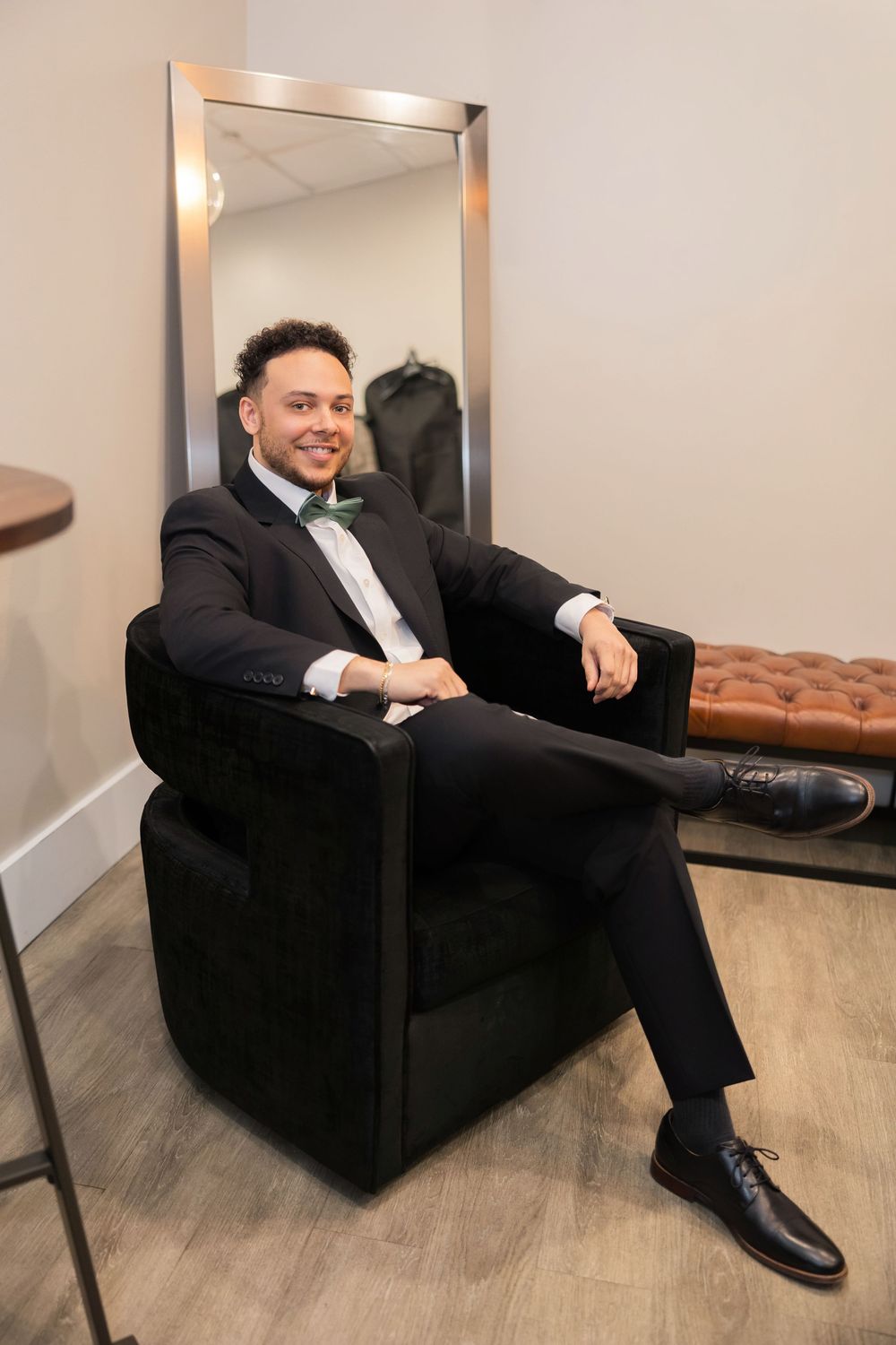Dapper groom in black tuxedo with green bow tie sits relaxed in velvet chair beside full-length mirror.