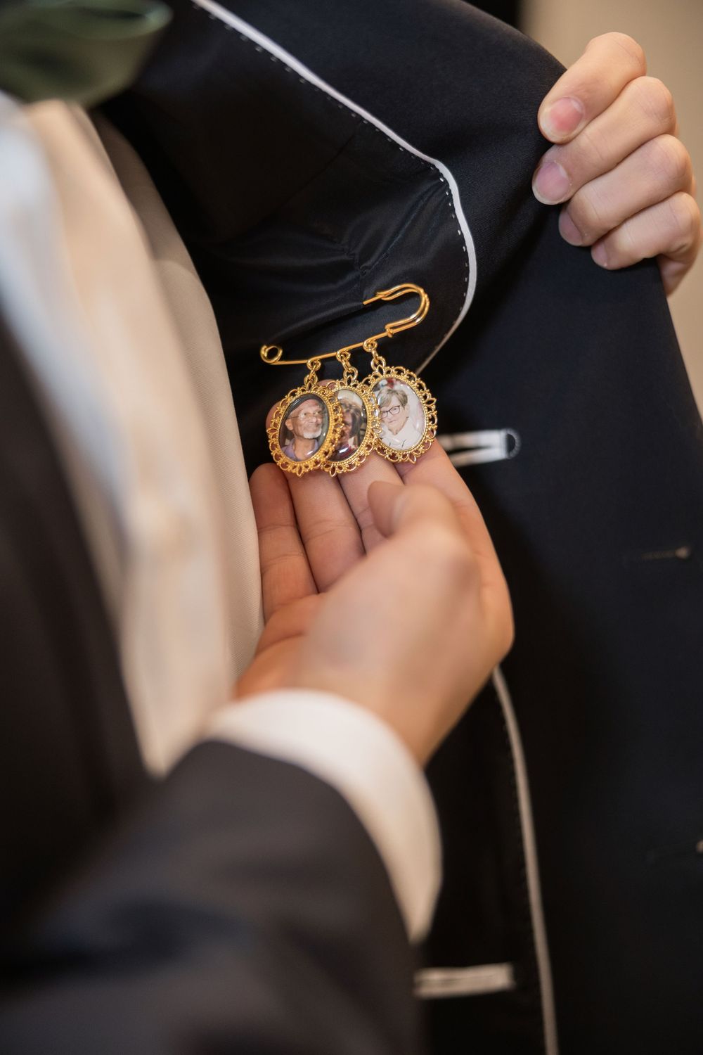 Groom pins sentimental gold photo locket memorial charm onto his dark suit jacket lapel.