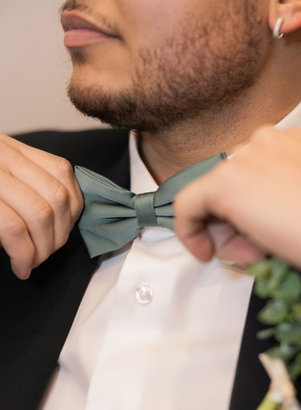 Groom adjusts sage green bow tie on white dress shirt while getting ready for wedding day.