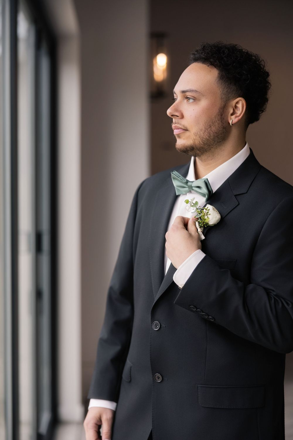 Handsome groom in black tuxedo adjusting boutonniere while gazing out window on wedding day.
