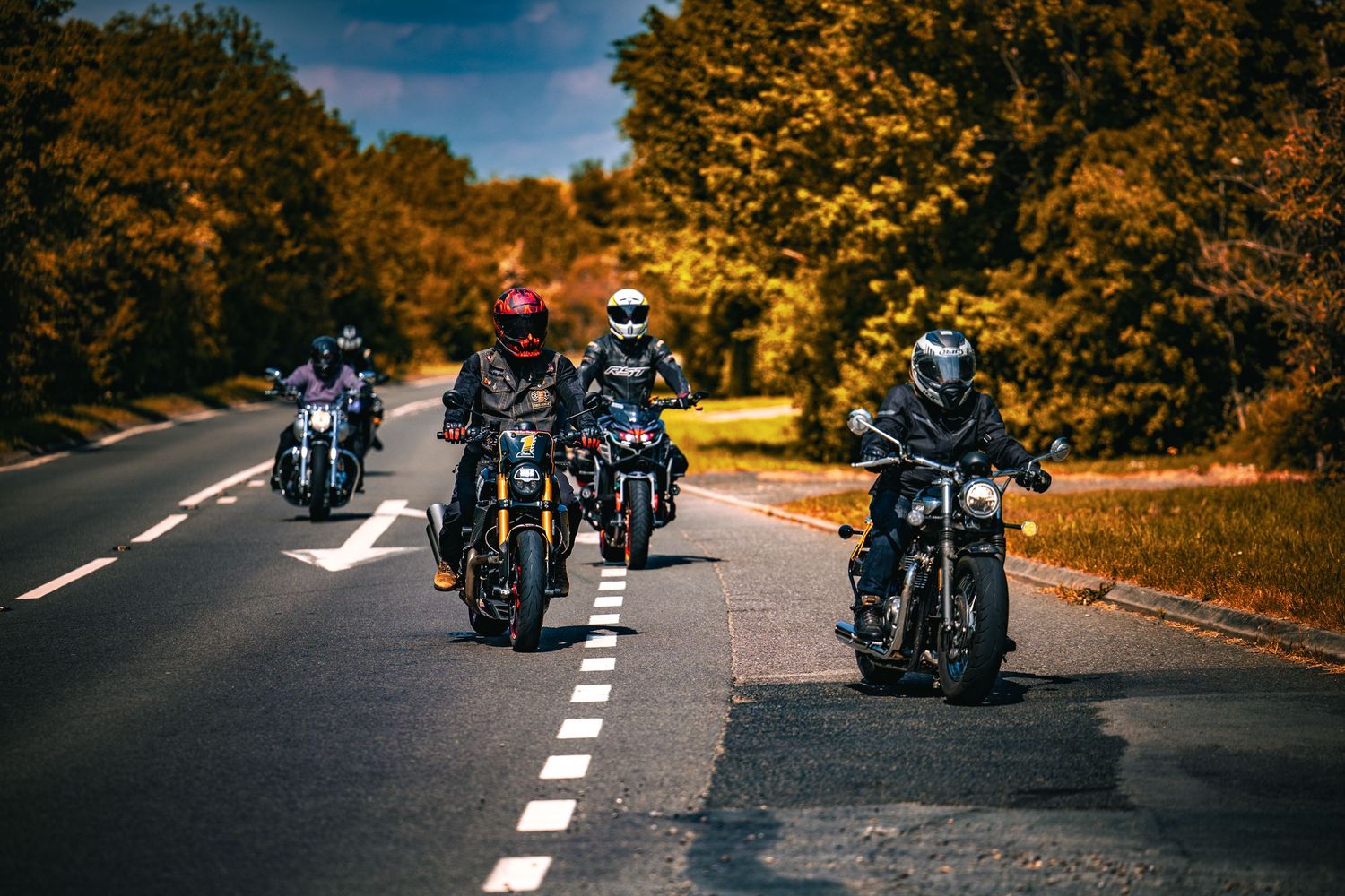 Group of motorcyclists riding in formation on a tree-lined road during a sunny autumn day.
