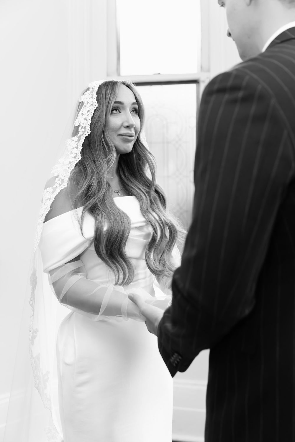 Black and white photo of a bride in an off-shoulder gown and lace veil holding hands with groom during wedding ceremony.