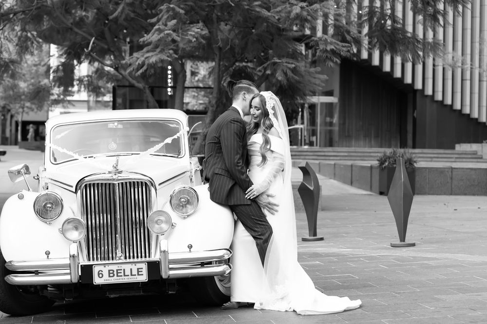 Bride and groom pose beside a vintage white Jaguar wedding car in a romantic black and white outdoor portrait.