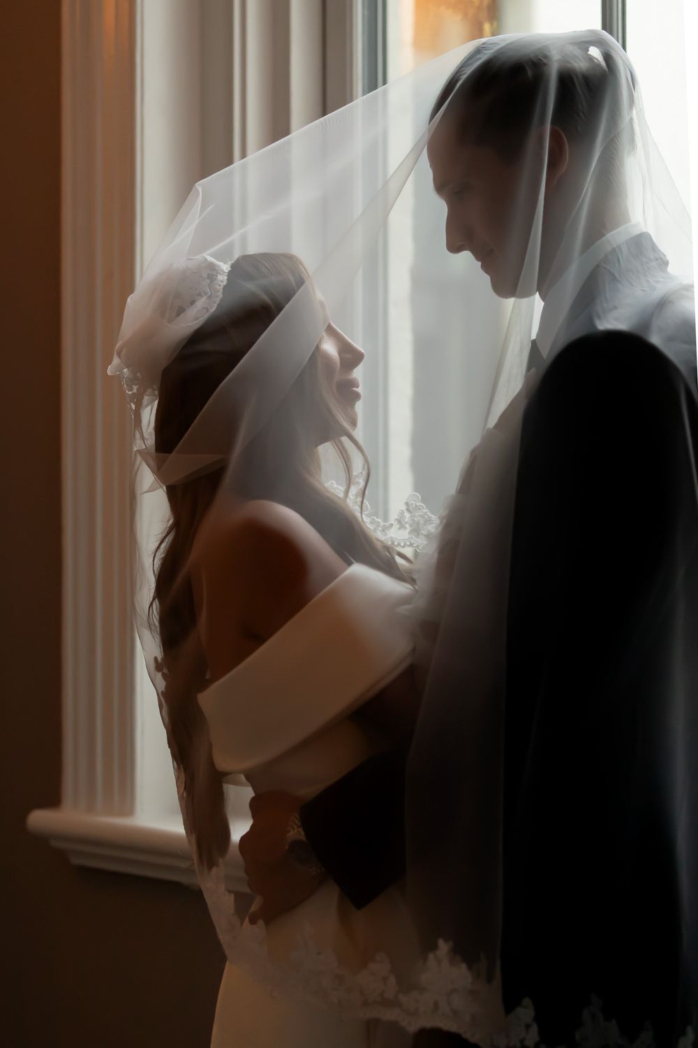 Bride and groom share an intimate moment under a wedding veil by a window in soft romantic lighting.