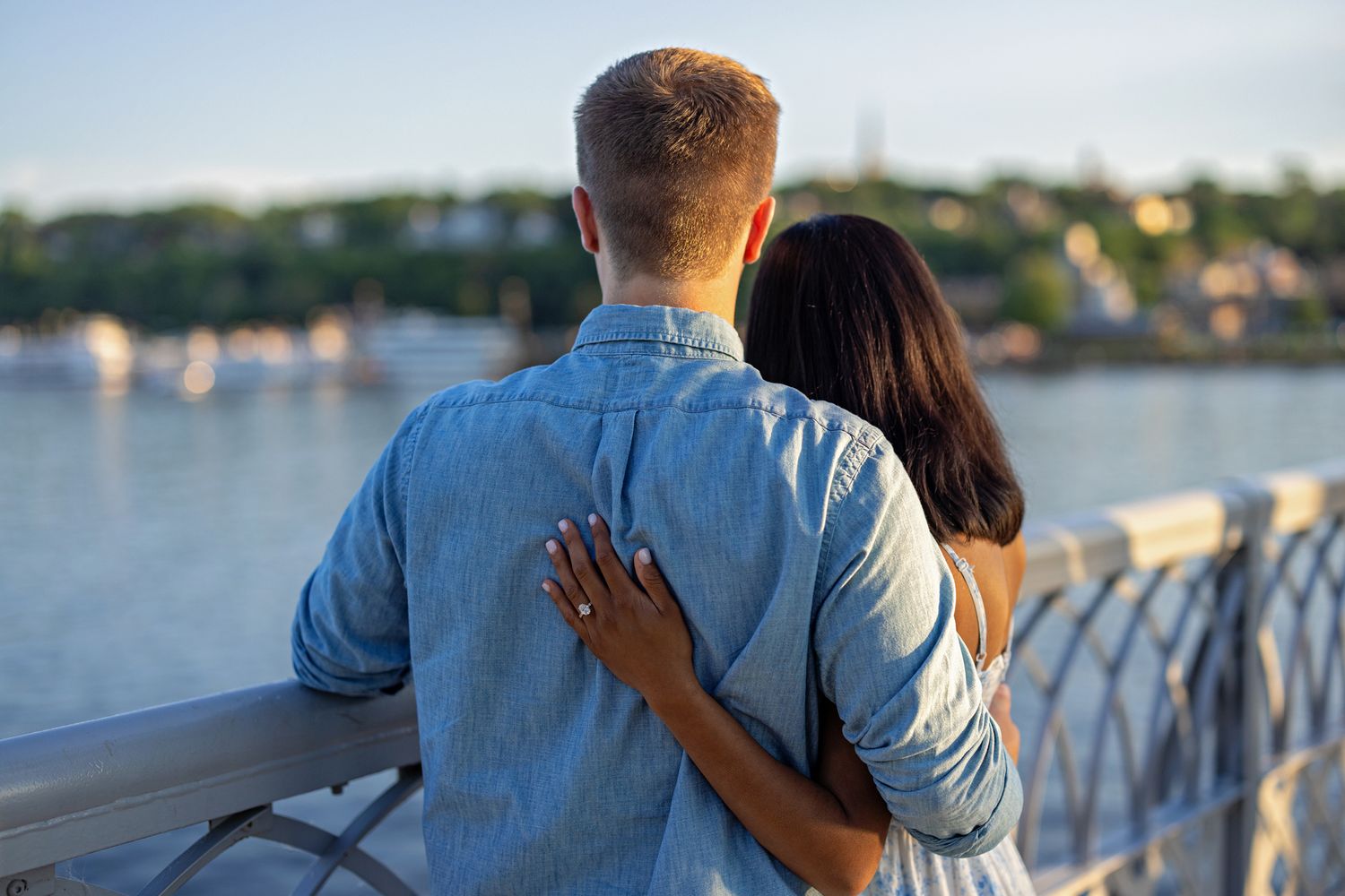 Romantic couple embracing on a bridge railing with scenic river and town visible in the background.