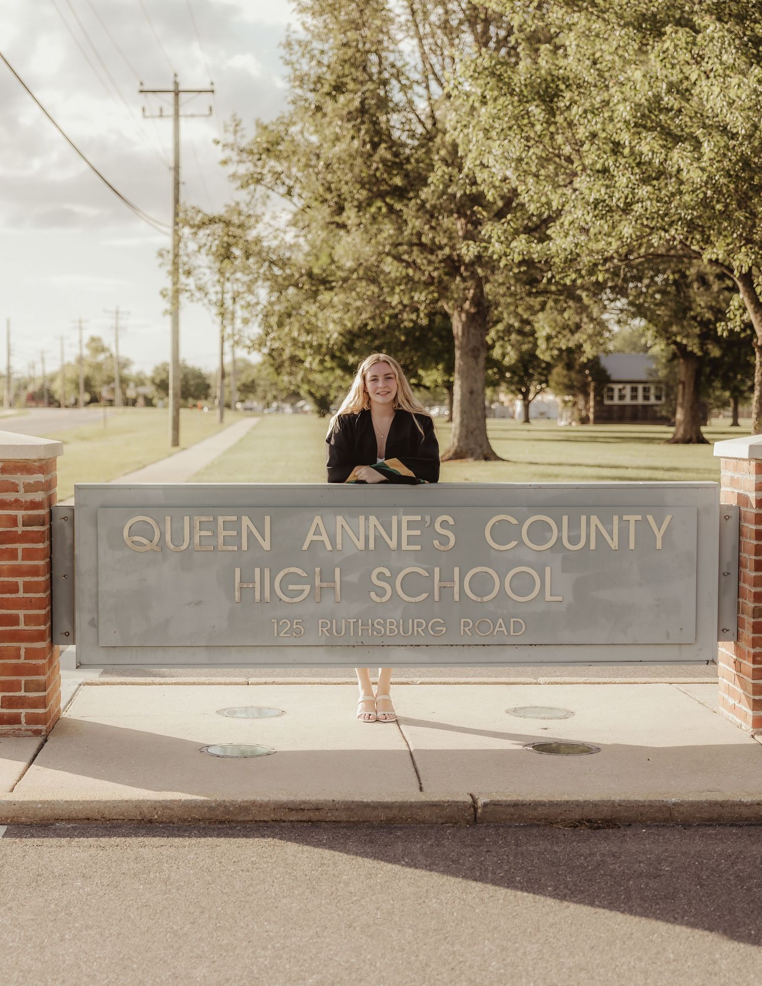 A student poses behind the Queen Annes County High School sign on Ruthsburg Road on a sunny day.