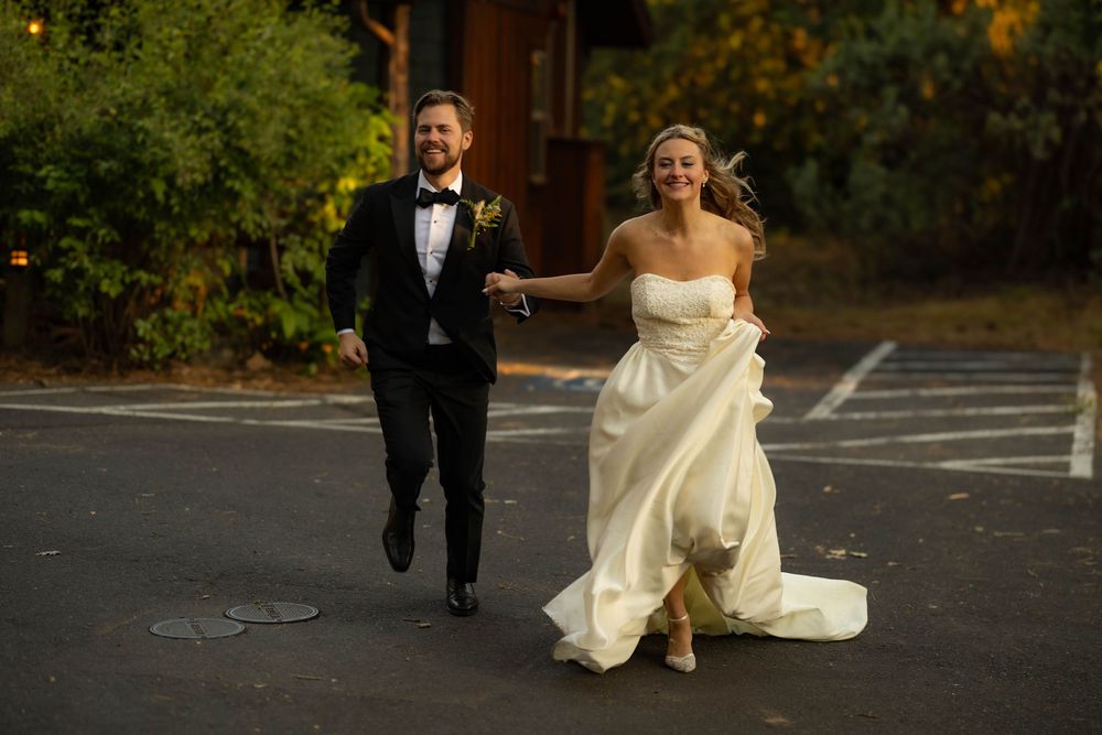 Smiling bride in white strapless gown and groom in black tuxedo run hand-in-hand across a parking lot.