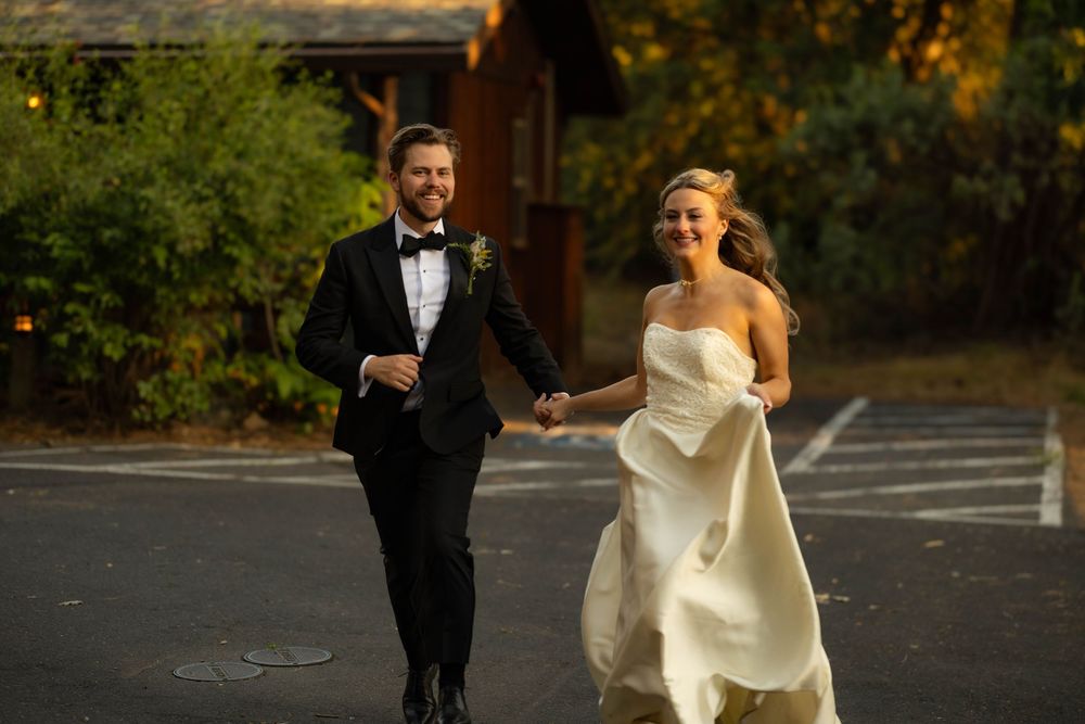Bride and groom run hand-in-hand laughing in parking lot surrounded by autumn foliage.