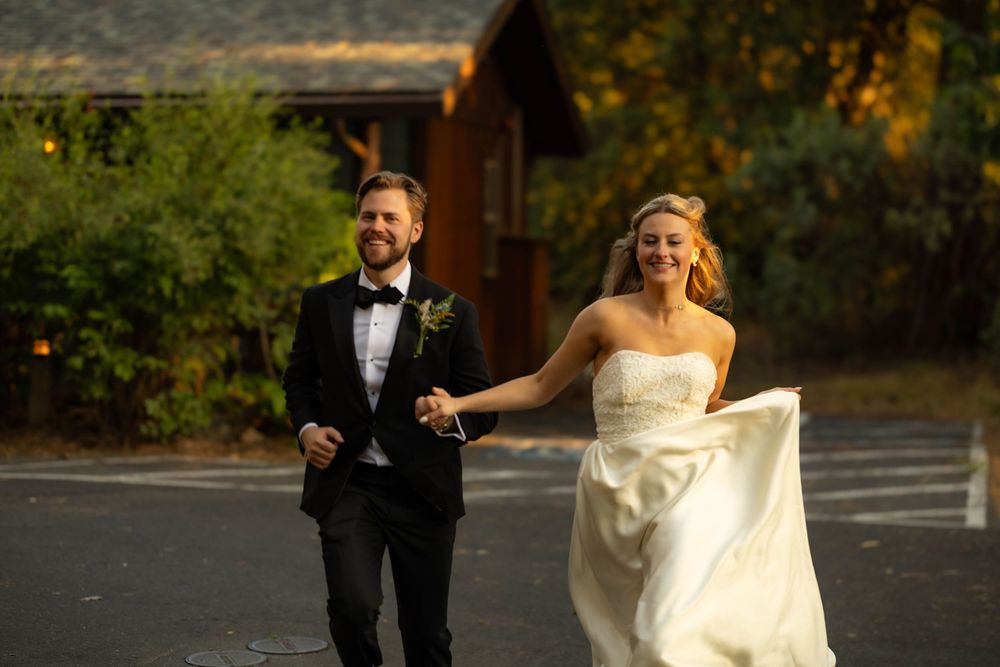 Joyful bride and groom walking hand in hand outdoors in golden afternoon sunlight near a cabin.