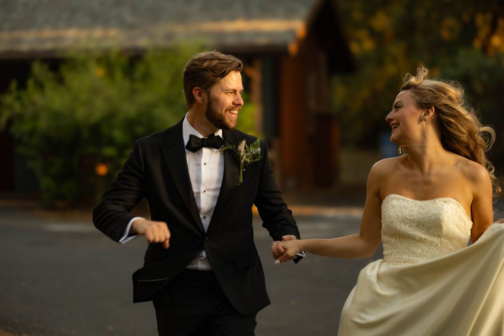 Joyful bride and groom in elegant attire hold hands laughing outdoors during golden hour wedding.