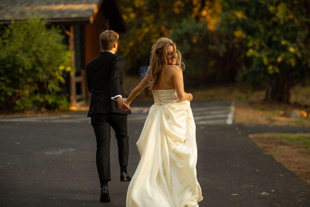 Bride and groom walk hand-in-hand down a sunlit driveway surrounded by golden autumn foliage.