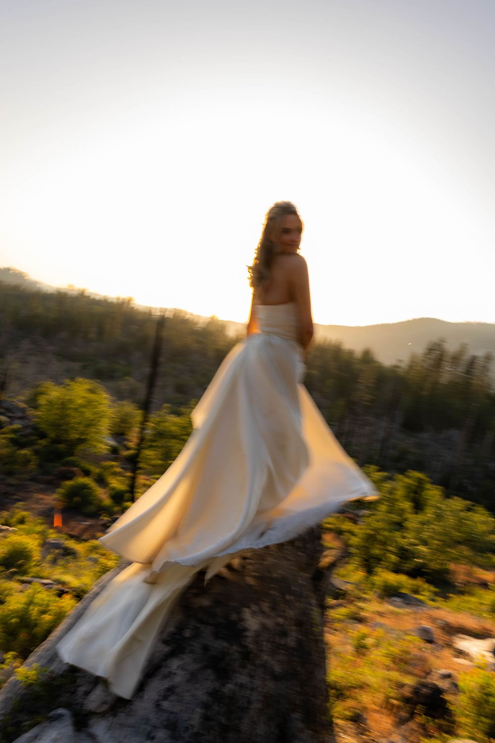 Bride in a flowing white gown standing on a rock at sunset with a mountain forest backdrop.