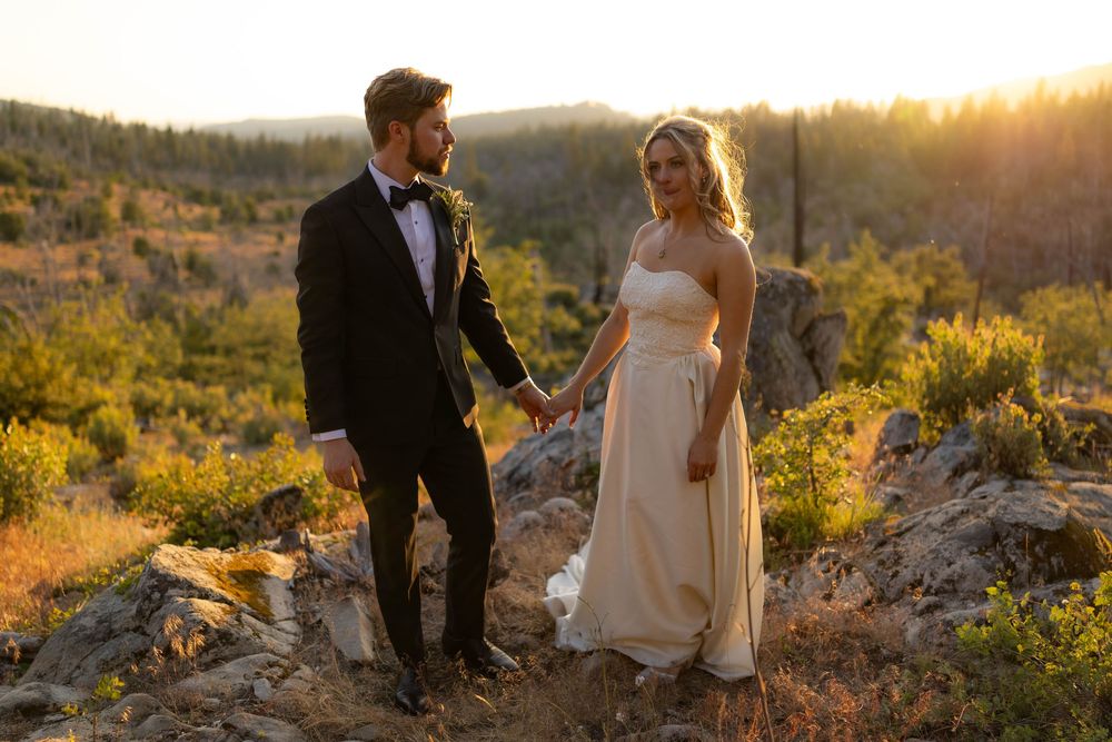 Bride in champagne gown and groom in black tuxedo hold hands on rocky terrain at golden sunset.