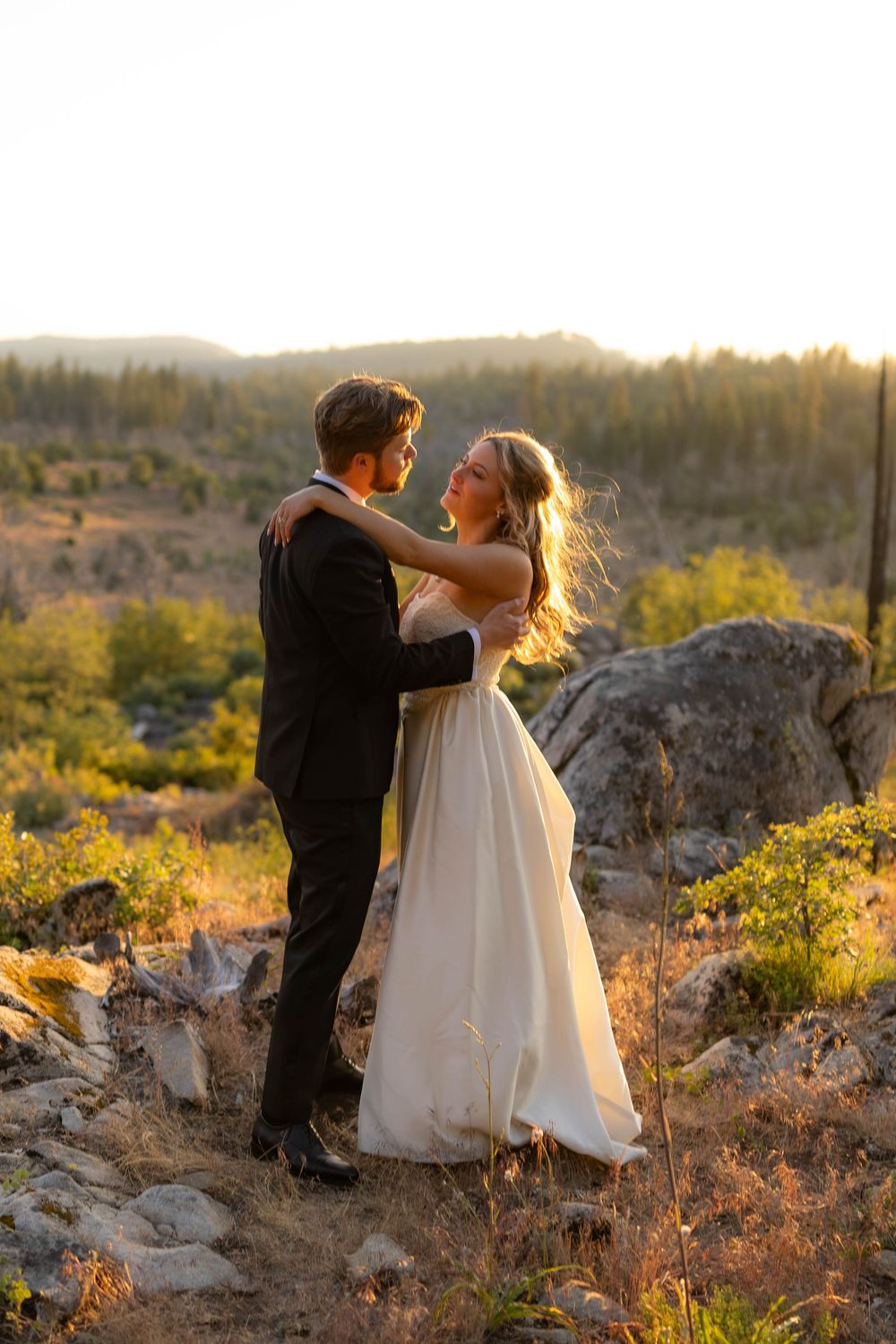 Bride and groom embrace at golden hour on rocky hillside, wearing wedding attire with forest backdrop.