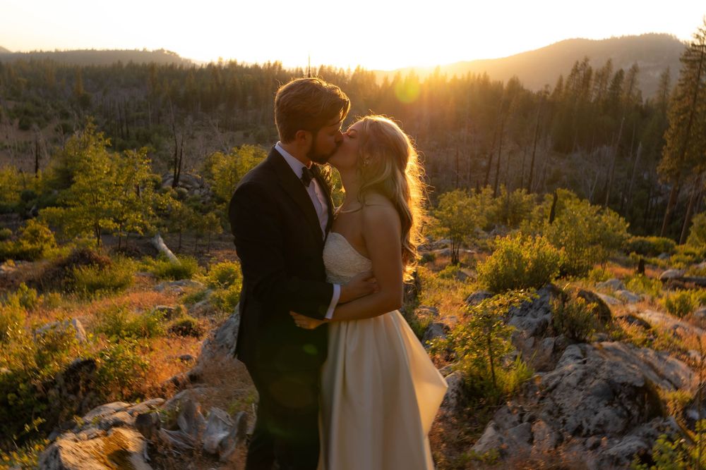 Bride and groom share a sunset kiss on rocky terrain with mountain forest backdrop.