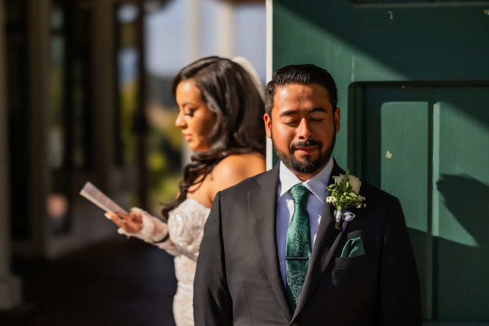 Groom in a grey suit leaning against a green wall while bride reads vows during first look.