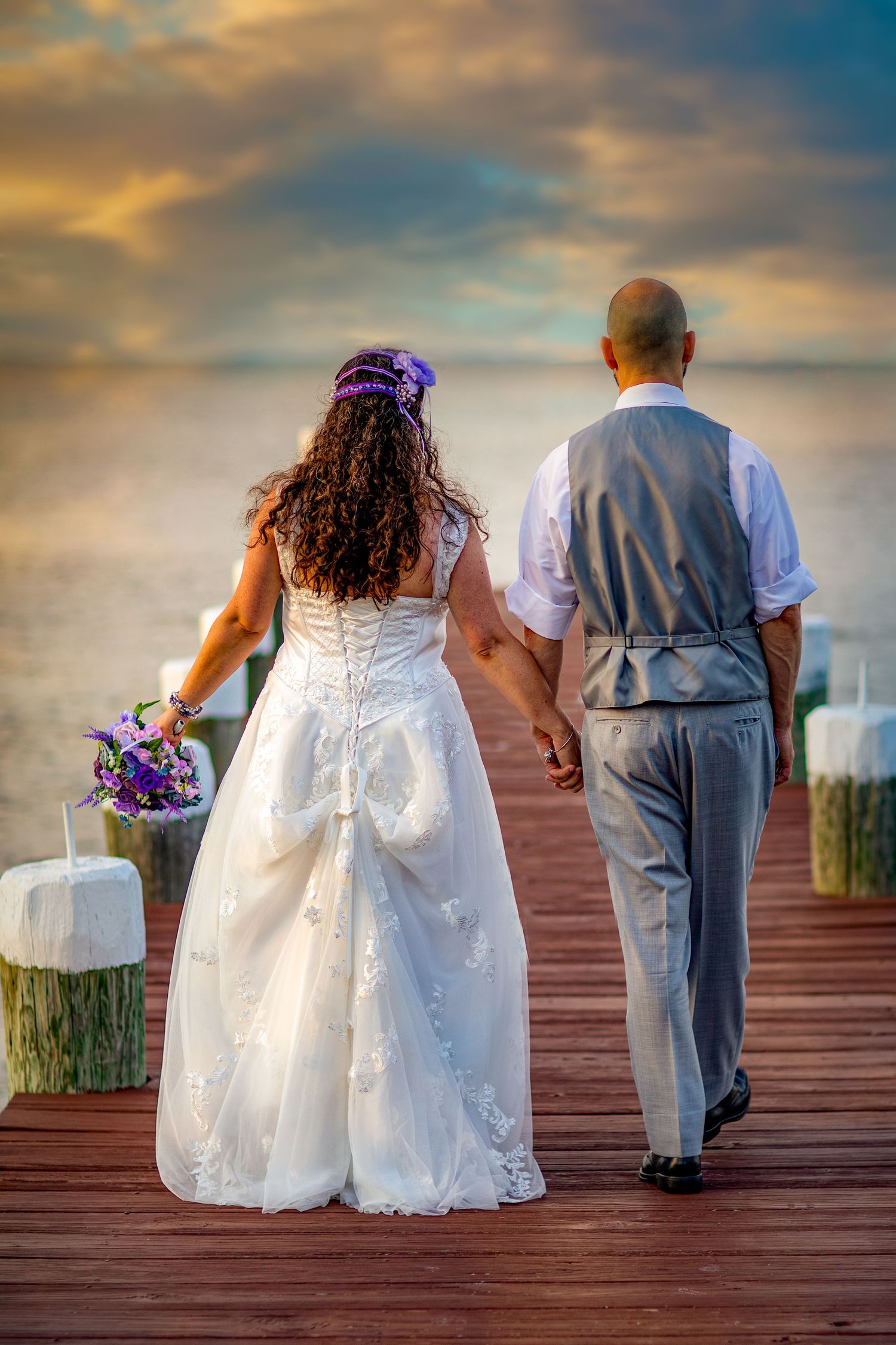Bride and groom holding hands walking on a wooden pier at sunset, bride in white lace gown holding purple bouquet.