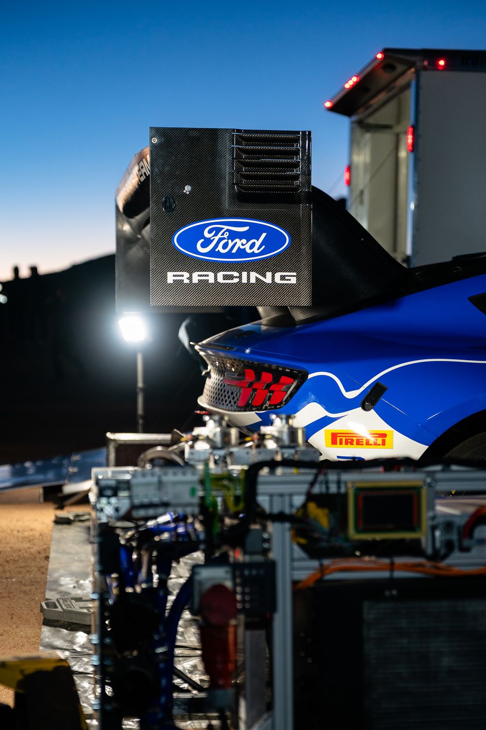 Ford Racing car on display at dusk with Pirelli tires, illuminated by bright lights against a clear blue sky.