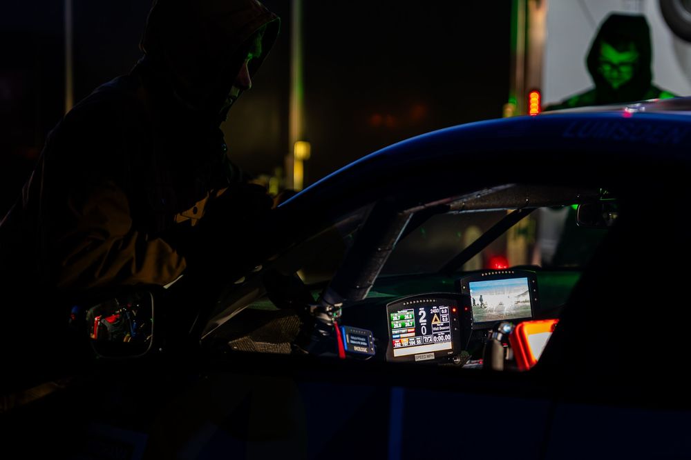 Dark racing car cockpit with illuminated dashboard screens and crew member visible in low light pit lane.