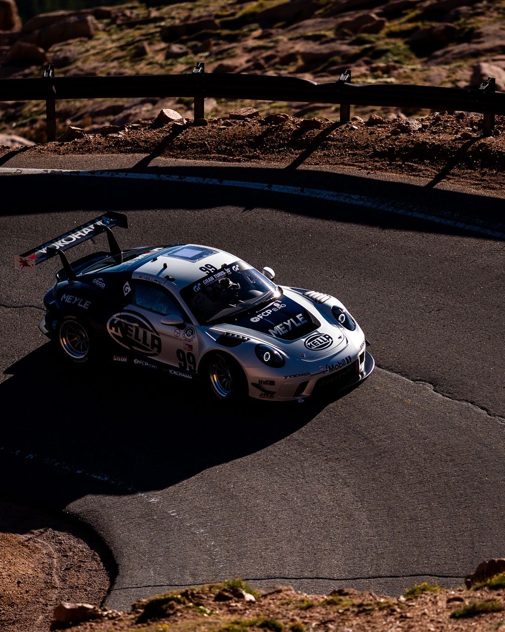 A silver race car with sponsorship decals navigates a sharp hairpin turn on a mountain racing circuit.