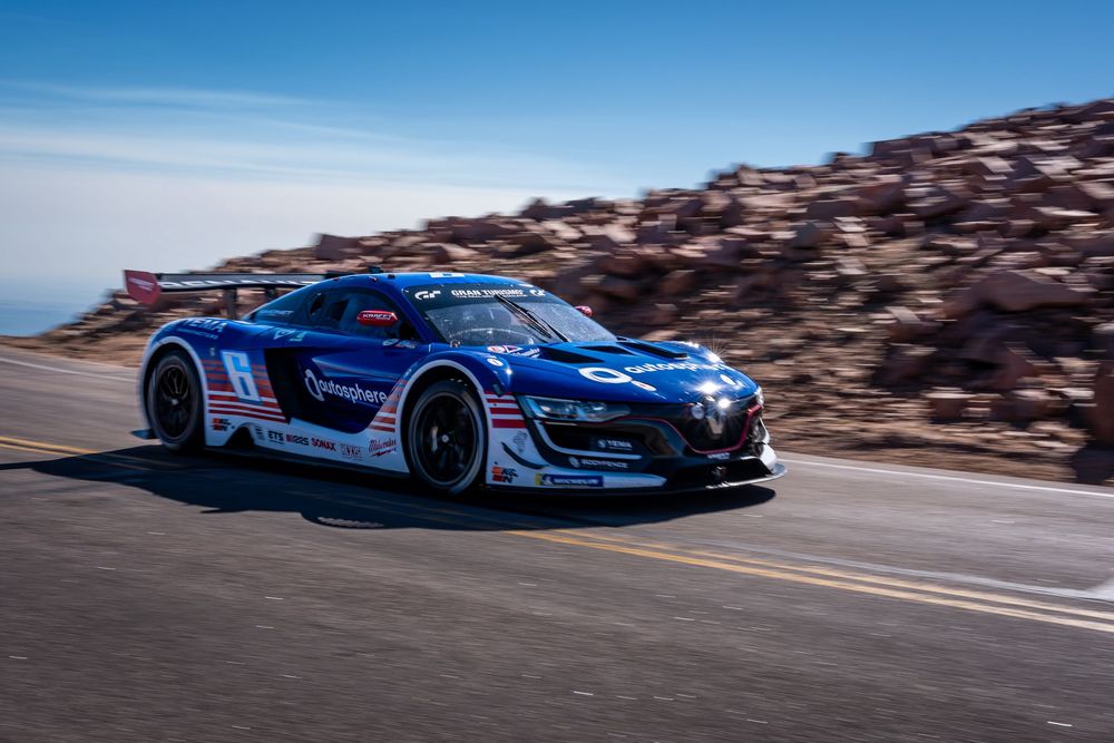 A blue racing car speeds along a mountain road at Pikes Peak International Hill Climb under a clear blue sky.