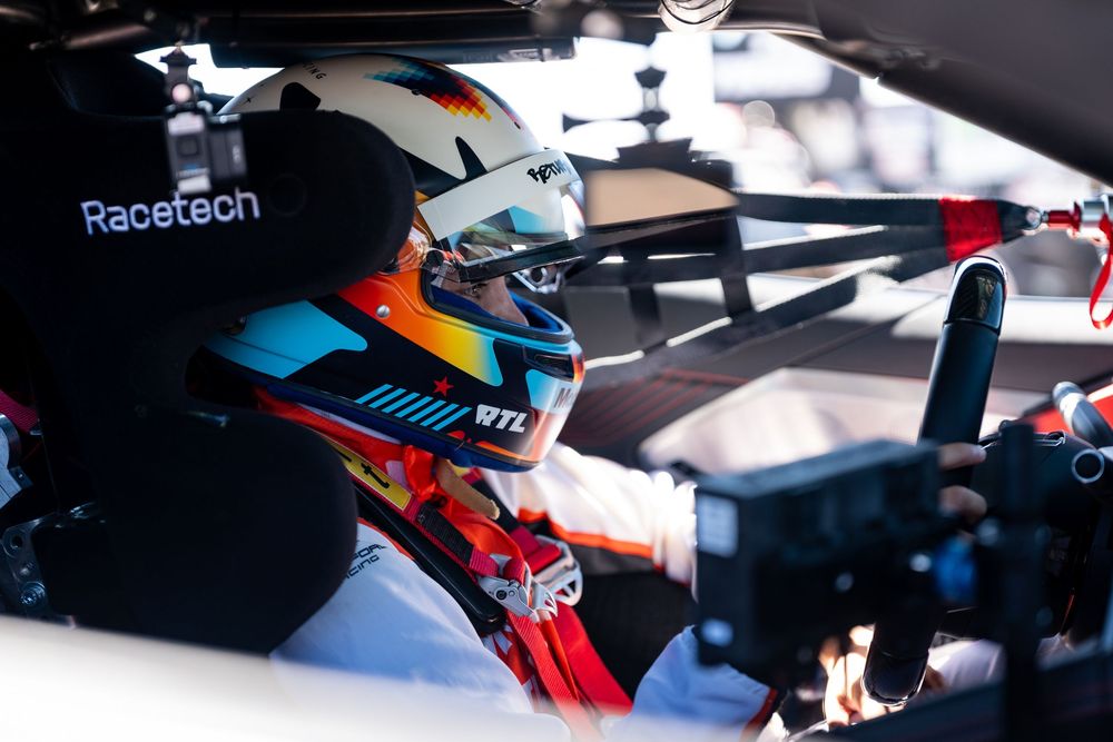 Racing driver in helmet and firesuit sits in cockpit of race car with Racetech sponsorship visible.