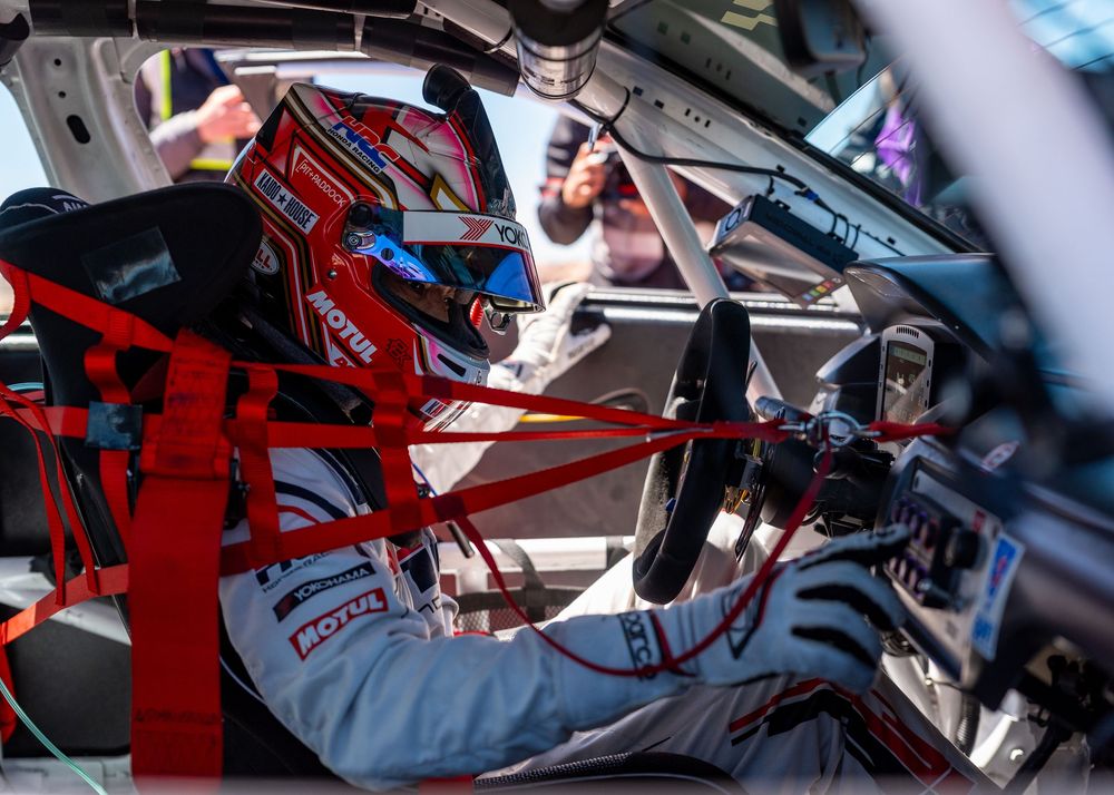 A race car driver in a helmet and red harness sits inside a racing car cockpit during a motorsport event.